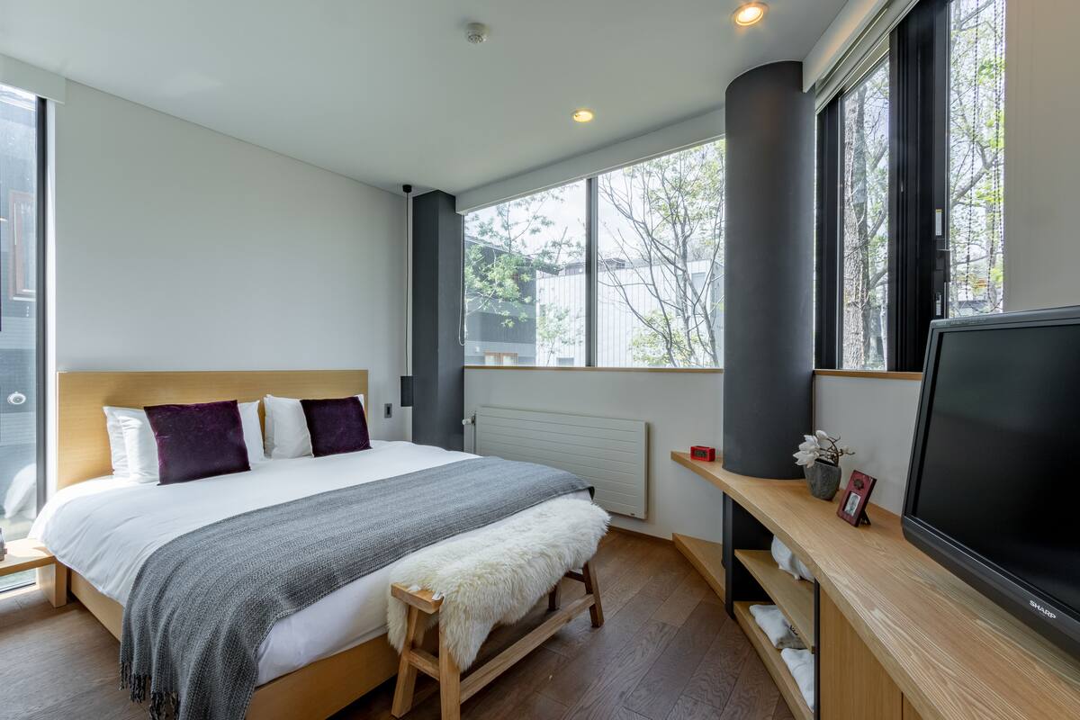 A modern bedroom with a large bed, a TV, and a window with a view of trees.
