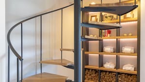 A spiral staircase with wooden steps and metal railings, leading to a lower level with a built-in shelf unit displaying towels and books.