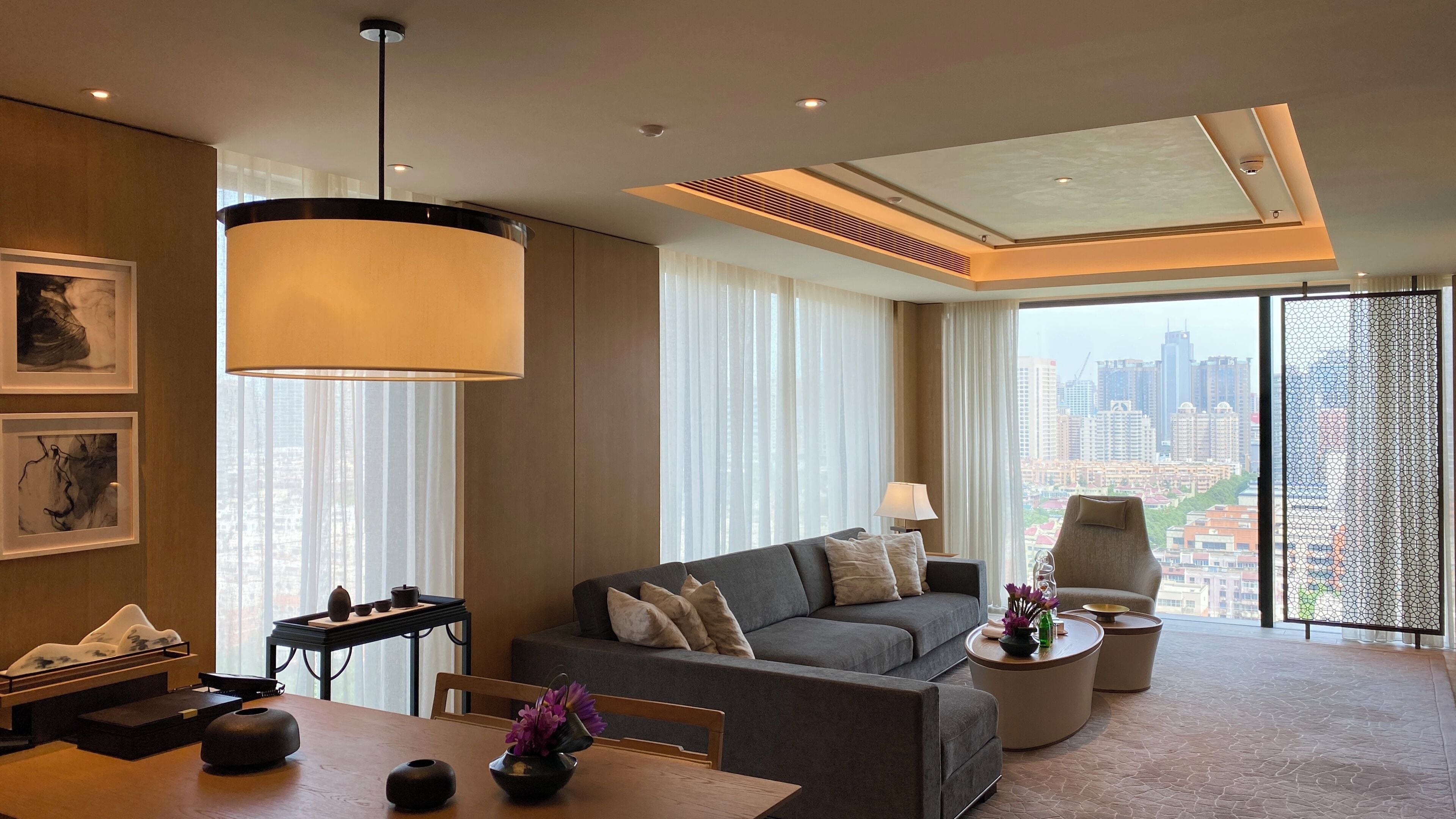 A modern hotel room with a sofa, a dining table, a chair, a lamp, and a view of the cityscape.