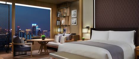Habitación de hotel con una cama grande, una zona de estar con vista a la ciudad, una mesita y un banco.