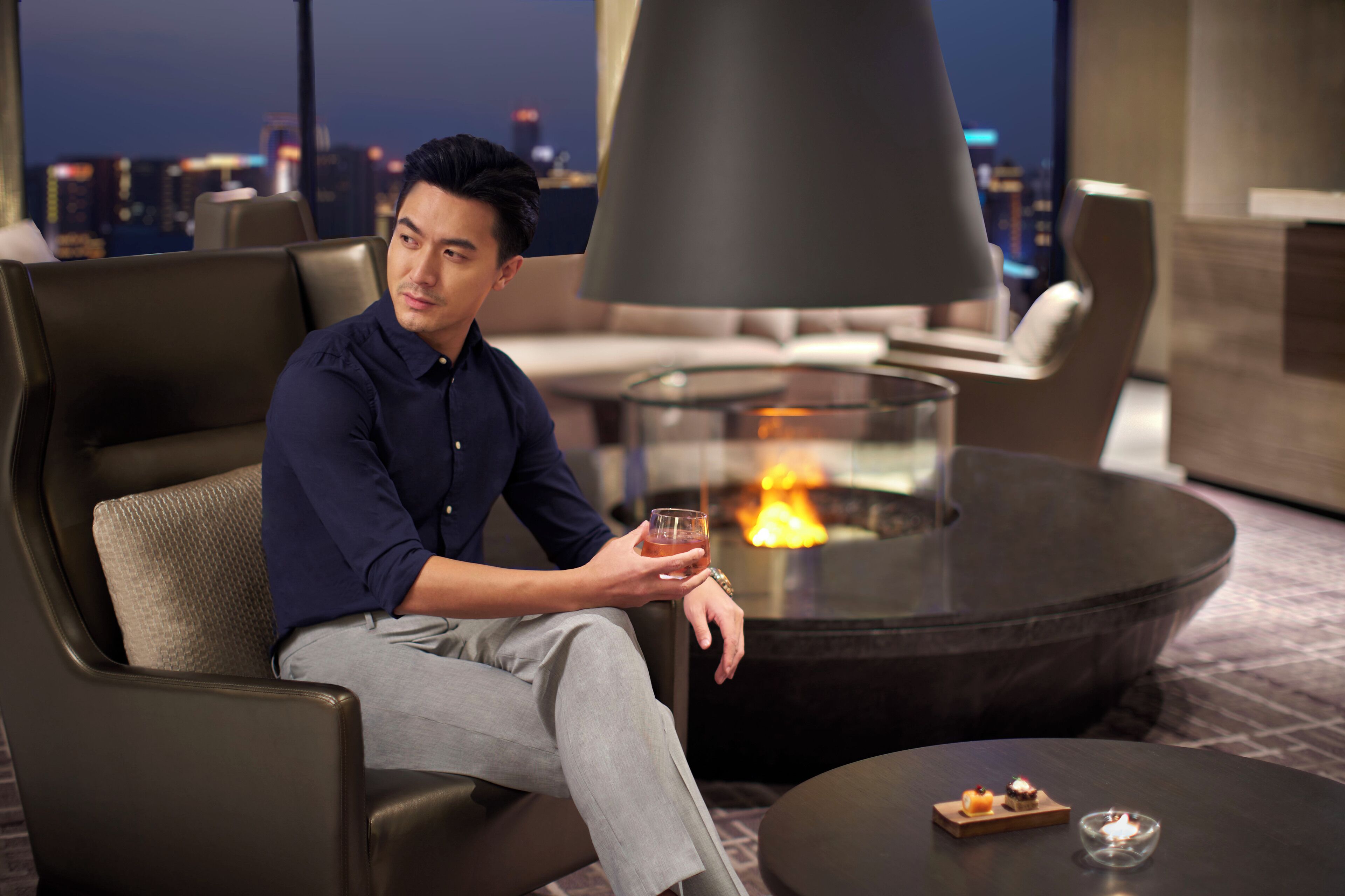 A man sitting on a leather sofa in a modern lounge area with a glass-top table and a small table with candles.