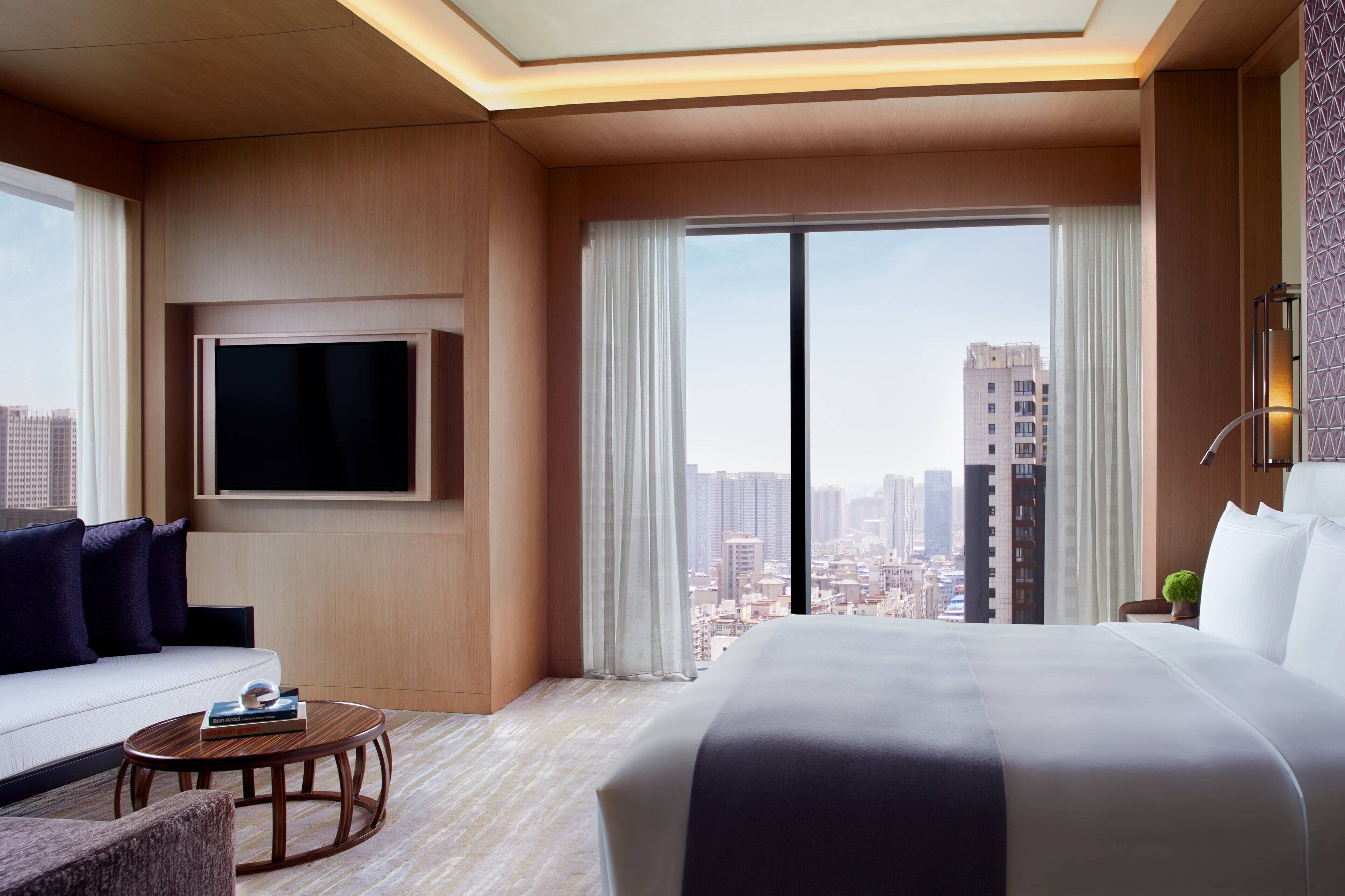 A modern hotel room with a large bed, a sofa, a TV mounted on the wall, and a city view through the window.