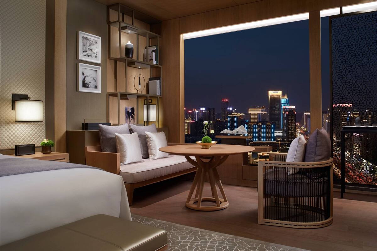 A modern hotel room with a large window offering a city skyline view, a cozy seating area with a sofa and armchair, and a small round table.