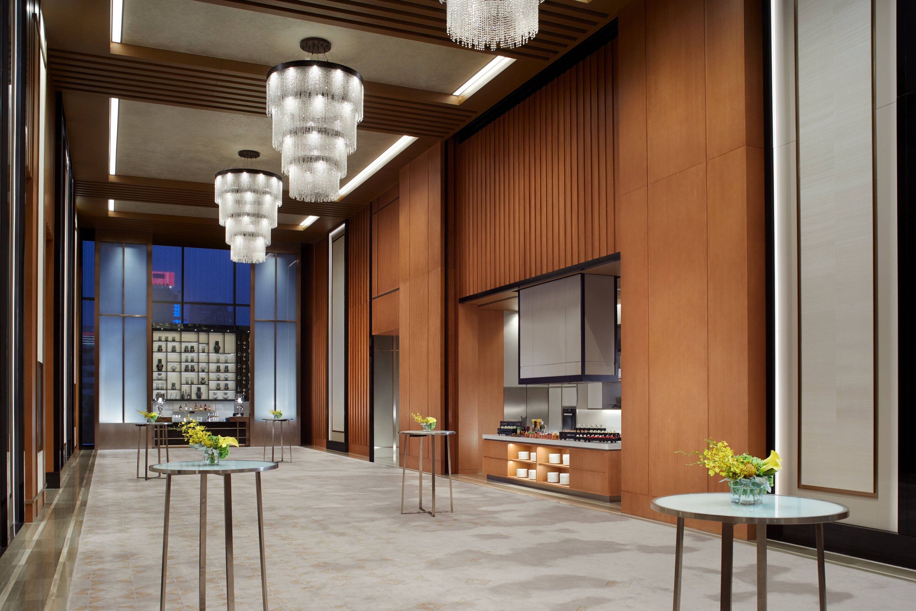 A spacious lobby with modern chandeliers, wooden paneling, and a view of a restaurant area.