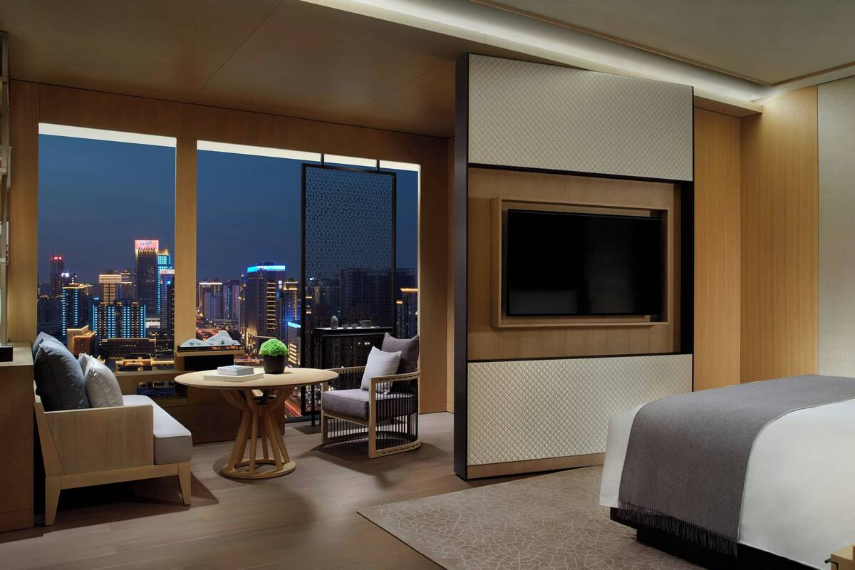 A modern hotel room with a city view, a flat-screen TV, a sofa, a chair, and a small table.