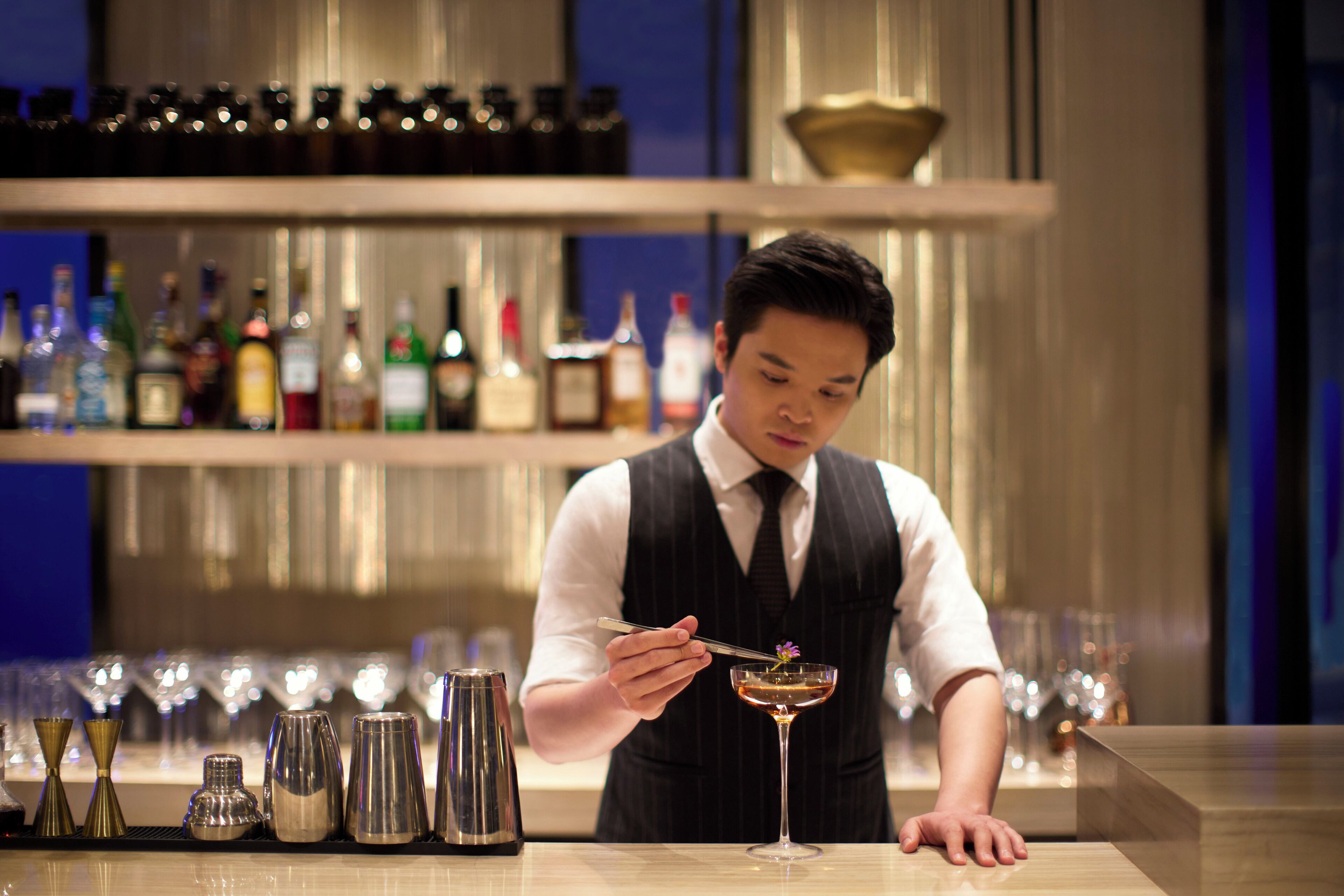 A bartender preparing a drink at a bar with various bottles and glasses in the background.