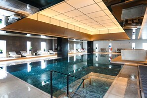 Une piscine intérieure moderne, avec des motifs géométriques en carreaux, des espaces de détente dotés de chaises blanches et un plafond haut avec des luminaires encastrés.