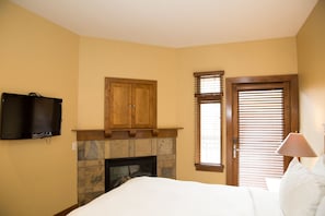 Quarto com cama, televisão na parede, armário de madeira, lareira de pedra e janela com persianas.