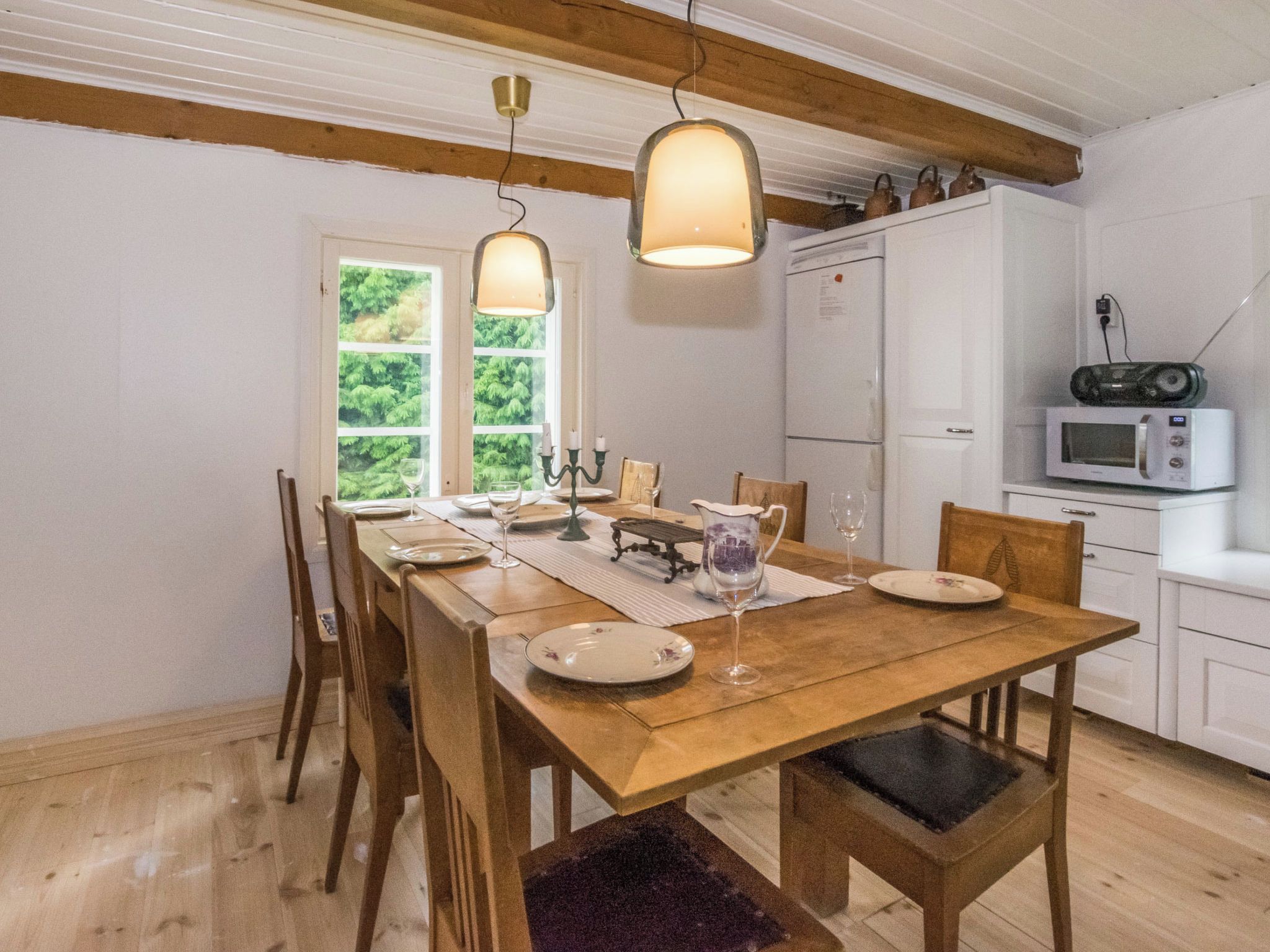 A dining area with a wooden table set for four, a window with a view of greenery, and a kitchen with a microwave and kettle.