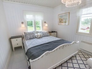 Une chambre à coucher avec un lit, deux tables de chevet, une commode, un lustre, une fenêtre donnant sur un beau paysage et un tableau encadré au mur.