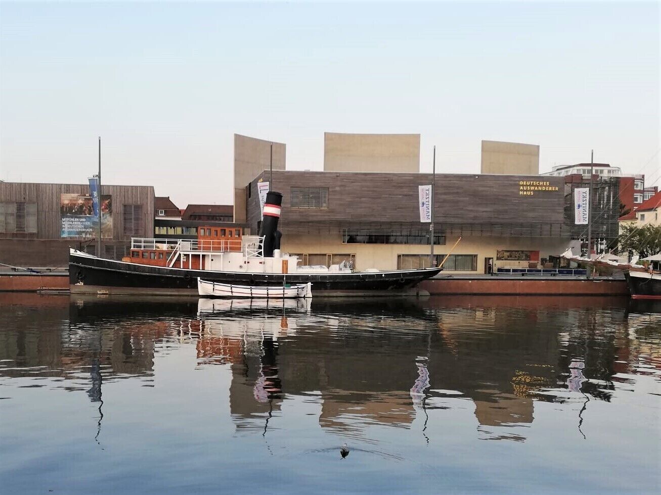 A calm river with two boats docked. In the background, there is a building with a sign that reads "DEUTSCHES AUSWANDERERHAUS".