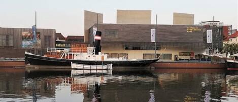A calm river with two boats docked. In the background, there is a building with a sign that reads "DEUTSCHES AUSWANDERERHAUS".
