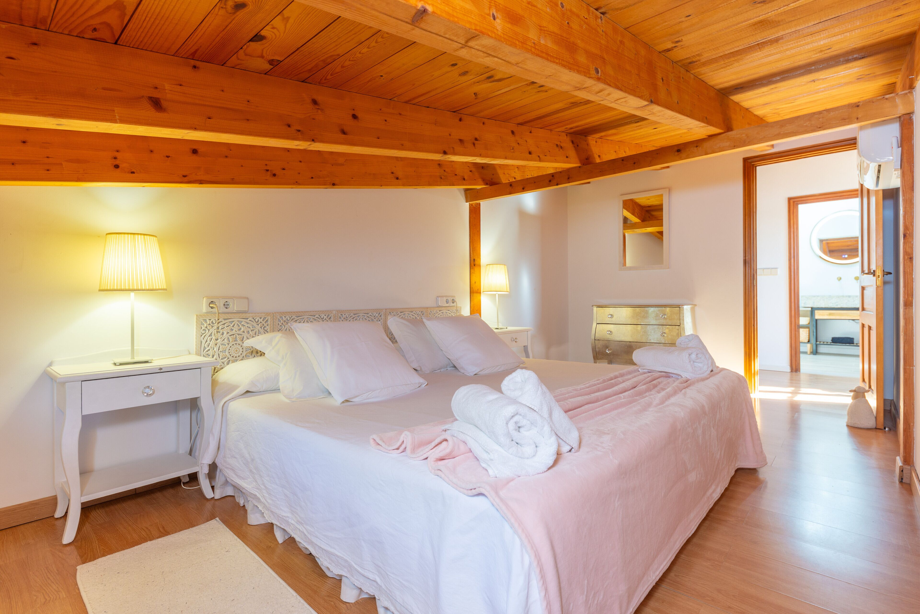 A bedroom with a wooden ceiling, a bed with white bedding, two bedside tables with lamps, and a white rug.