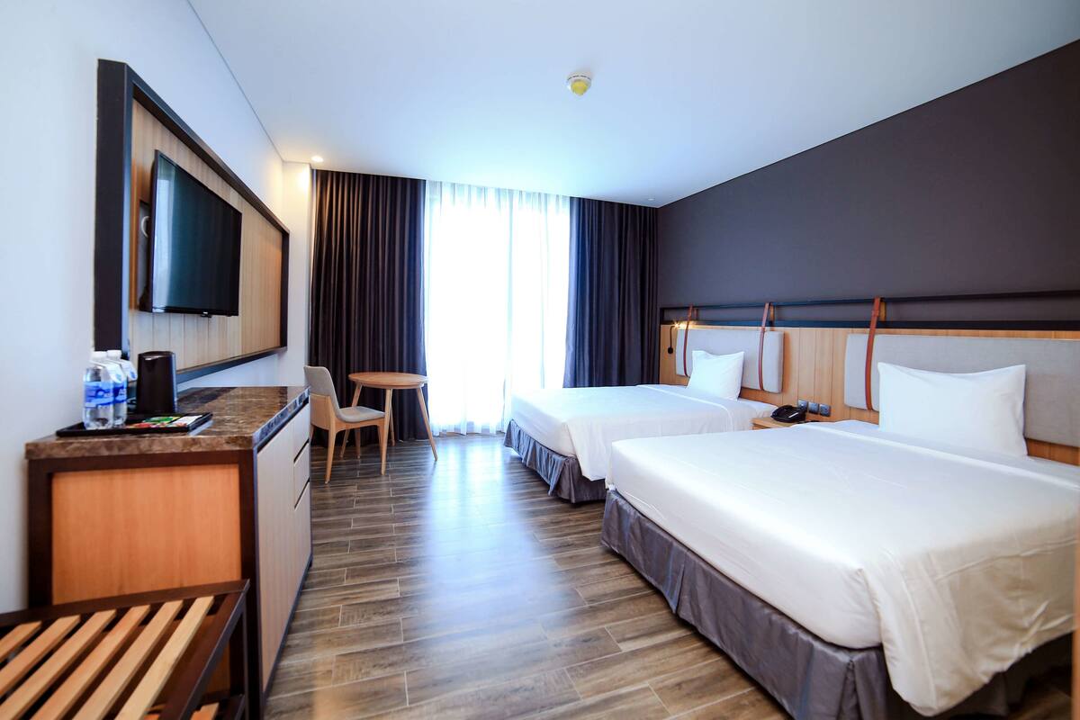 A hotel room with two beds, a desk, a TV, and a window with curtains.