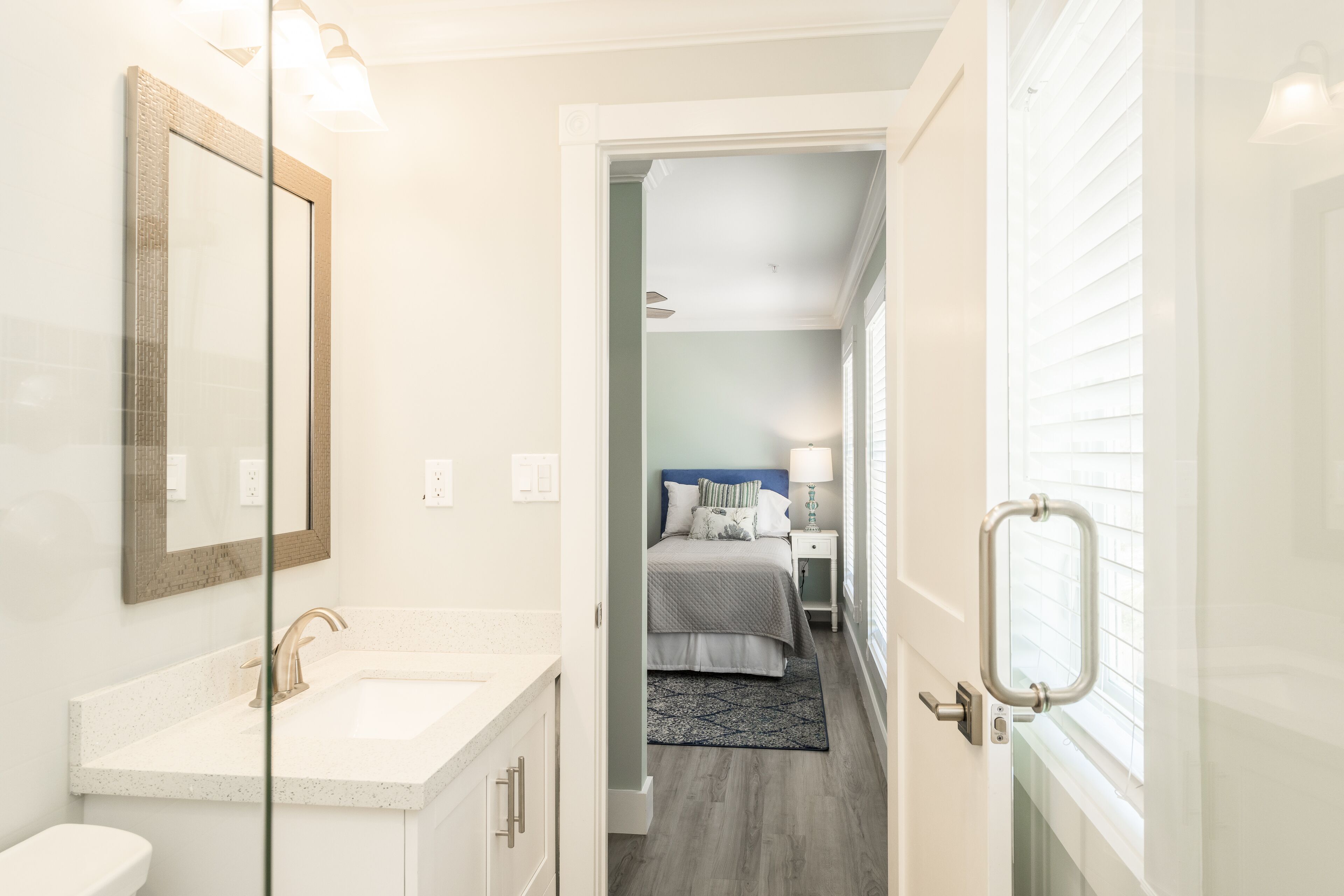 Une salle de bain avec un lavabo blanc, un miroir et une porte donnant sur une chambre à coucher équipée d’un lit et d’une table de chevet.
