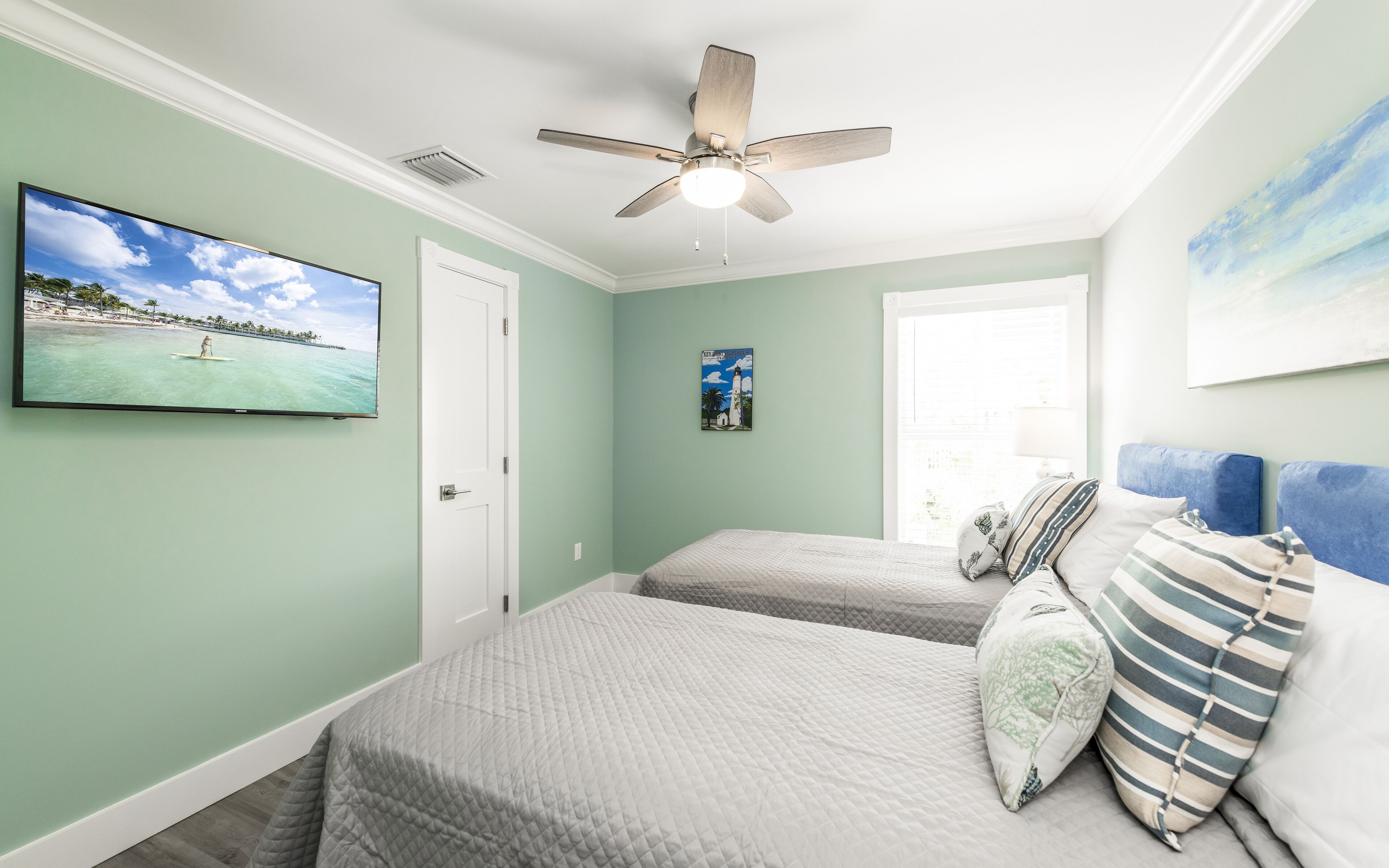 A bedroom with two beds, a ceiling fan, a television, and a window with a view of the beach.
