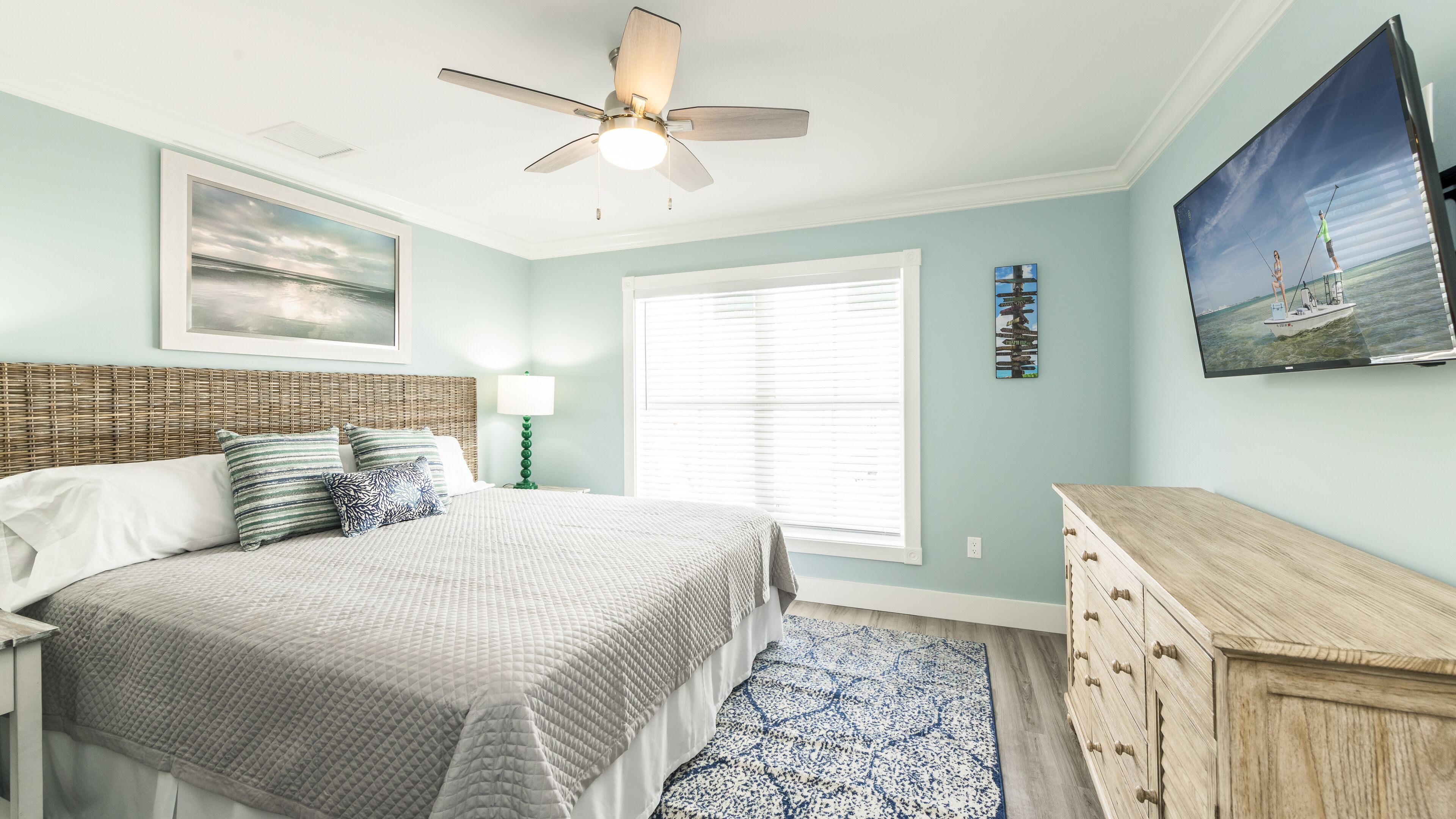 A bedroom with a bed, a ceiling fan, a window with blinds, a dresser, and a painting on the wall.