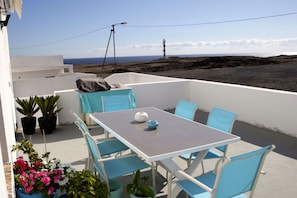 Um terraço com mesa e cadeiras, plantas em vasos e vista para o mar e um farol.
