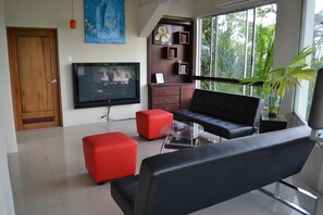 Una sala de estar moderna con un sofá negro, un reposapiés rojo y un televisor en la pared.