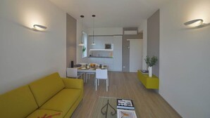Una sala de estar moderna con un sofá amarillo, un comedor con mesa y sillas blancas, y una cocina al fondo.