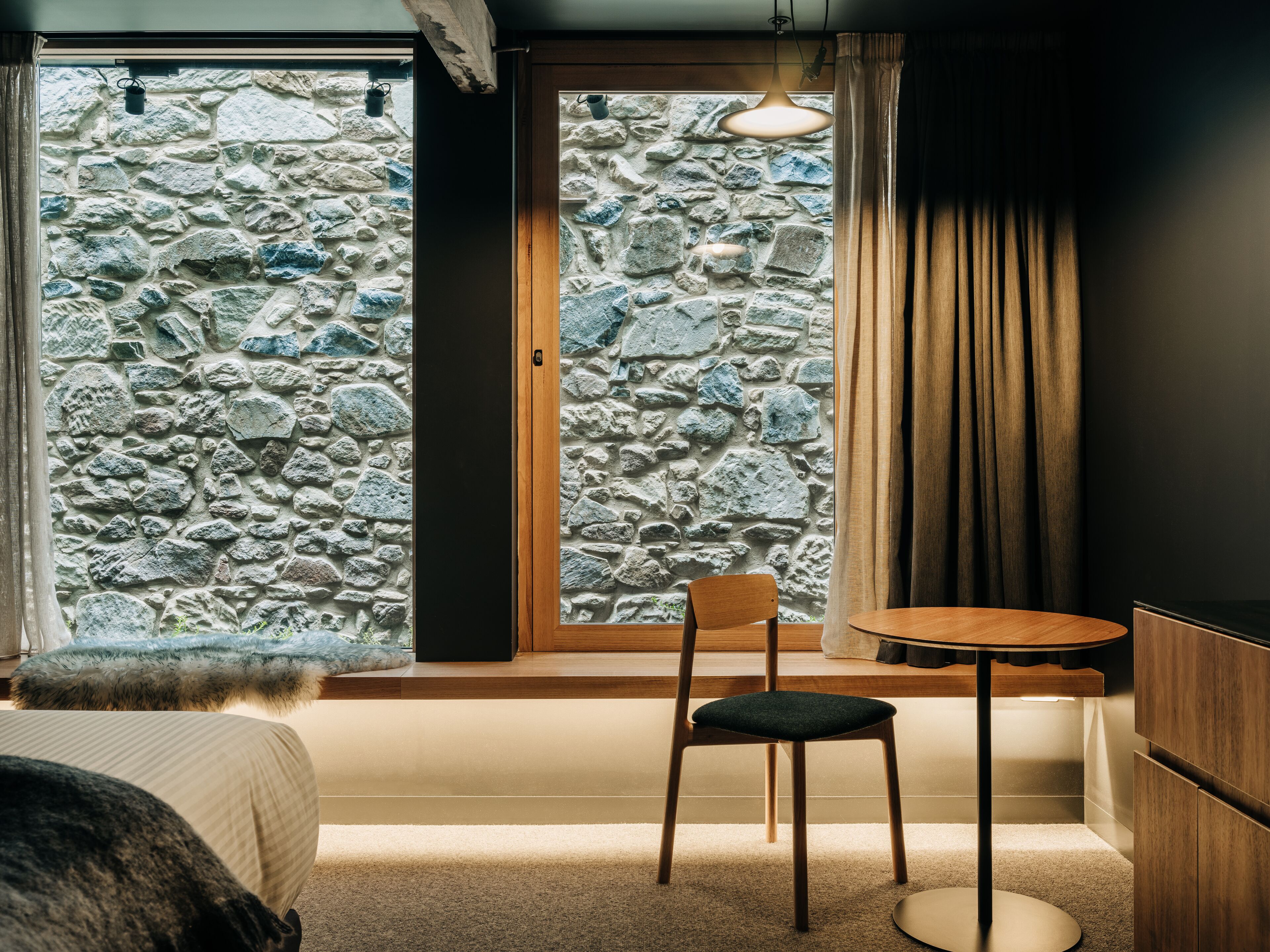 Una habitación con pared de piedra, una silla de madera, una pequeña mesa redonda y una cama con una manta de piel.