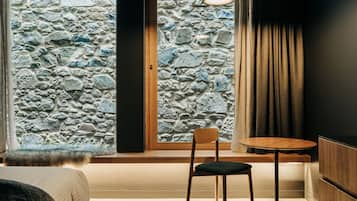 Una habitación con pared de piedra, una silla de madera, una pequeña mesa redonda y una cama con una manta de piel.