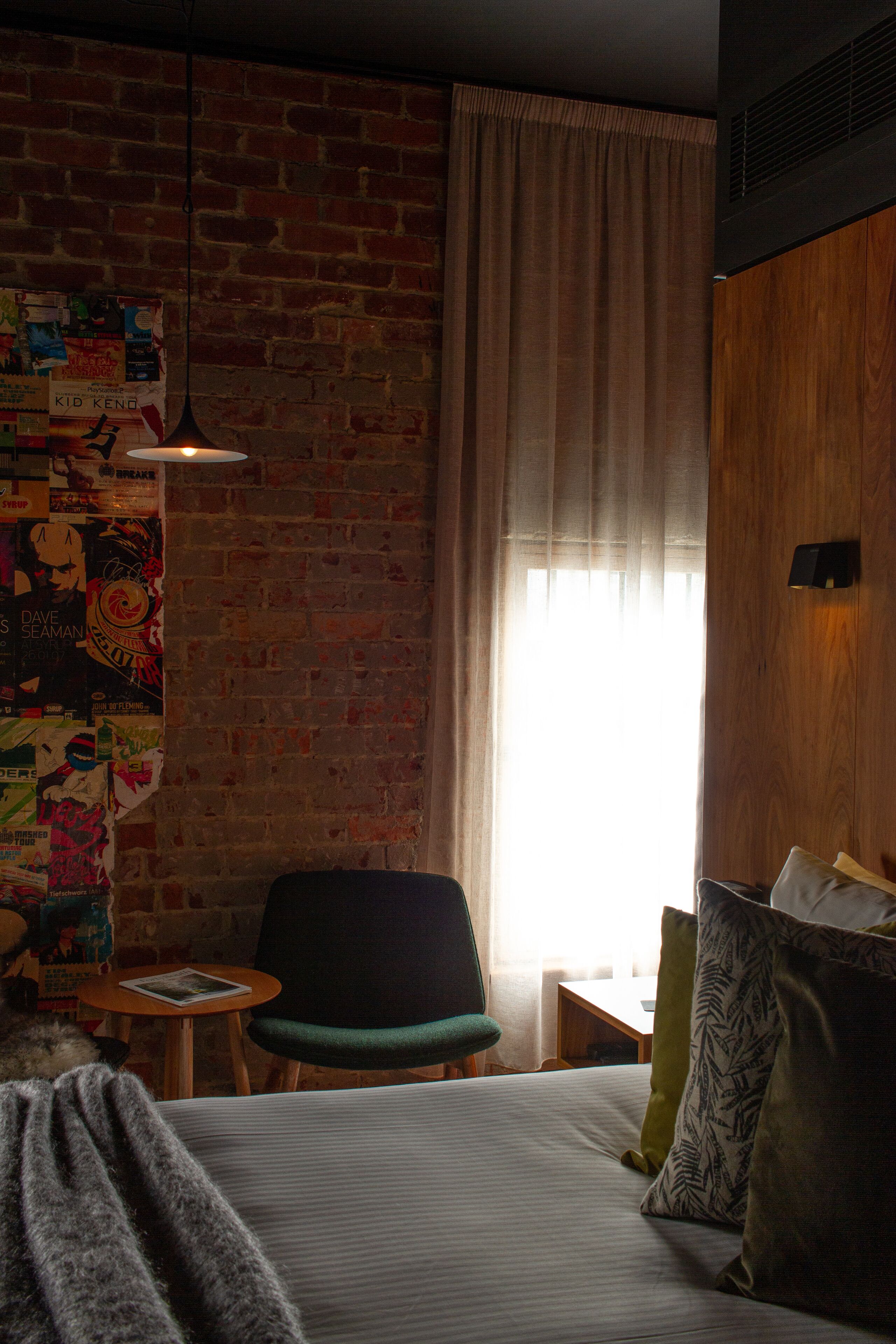 A room with a brick wall, a bed with pillows, a chair, a small table, and a hanging light fixture.