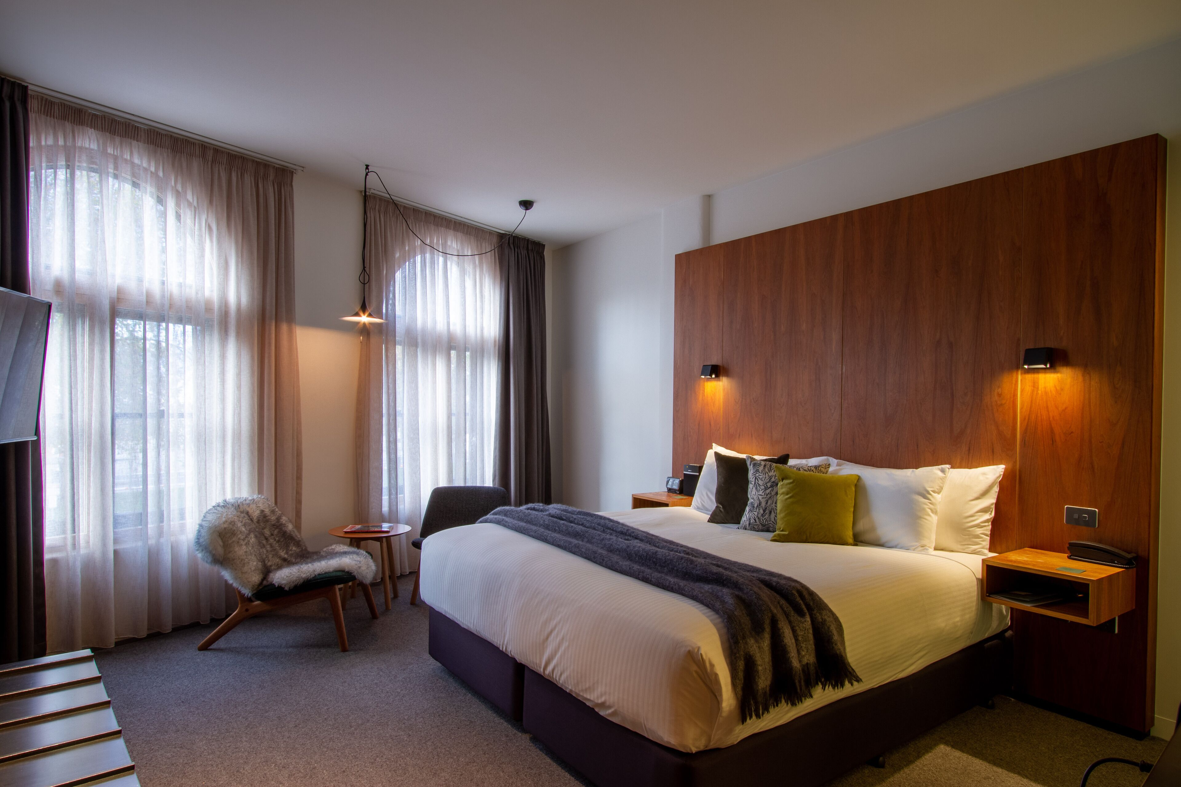 A modern hotel room with a large bed, a wooden headboard, a desk, and a chair.