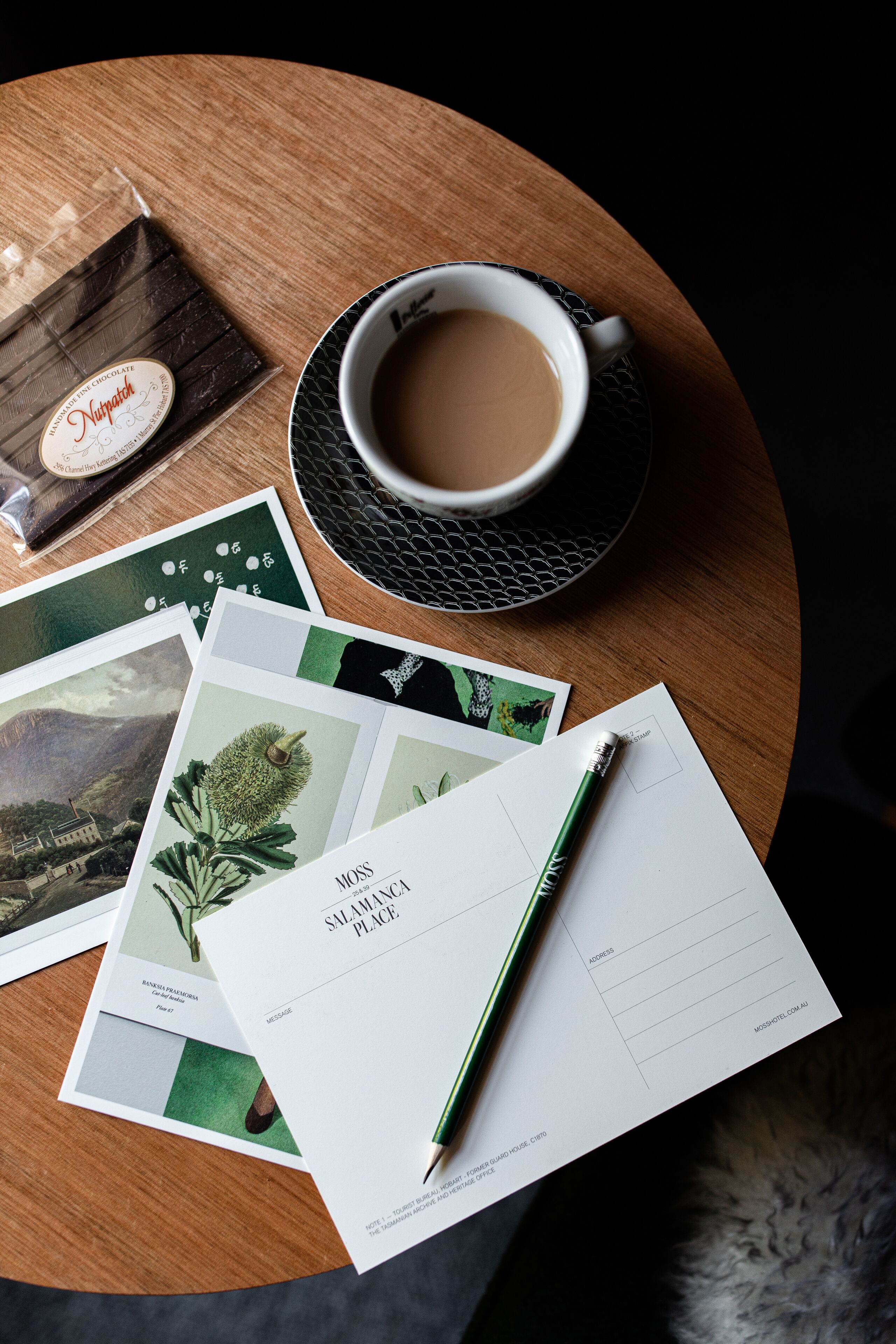 A wooden table with a cup of coffee, a chocolate bar, postcards, and a pen.