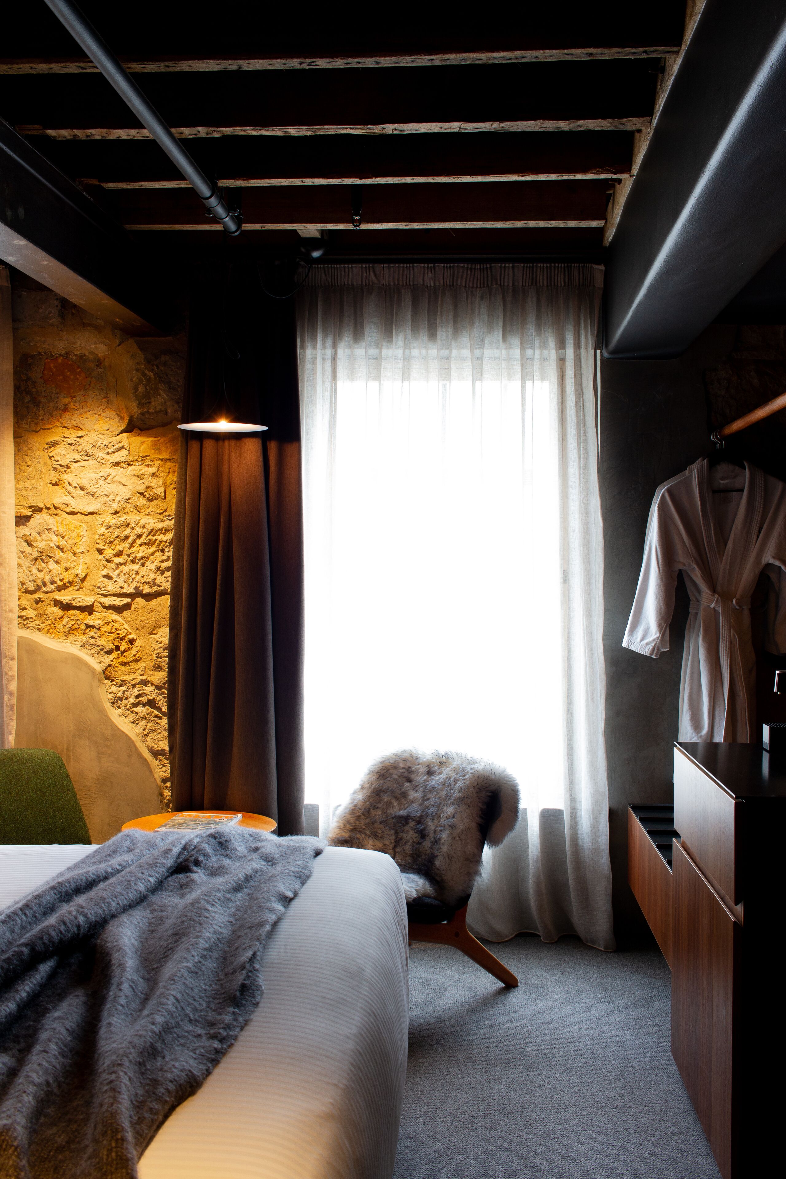 A hotel room with a bed, a chair, a robe hanging, and a stone wall.