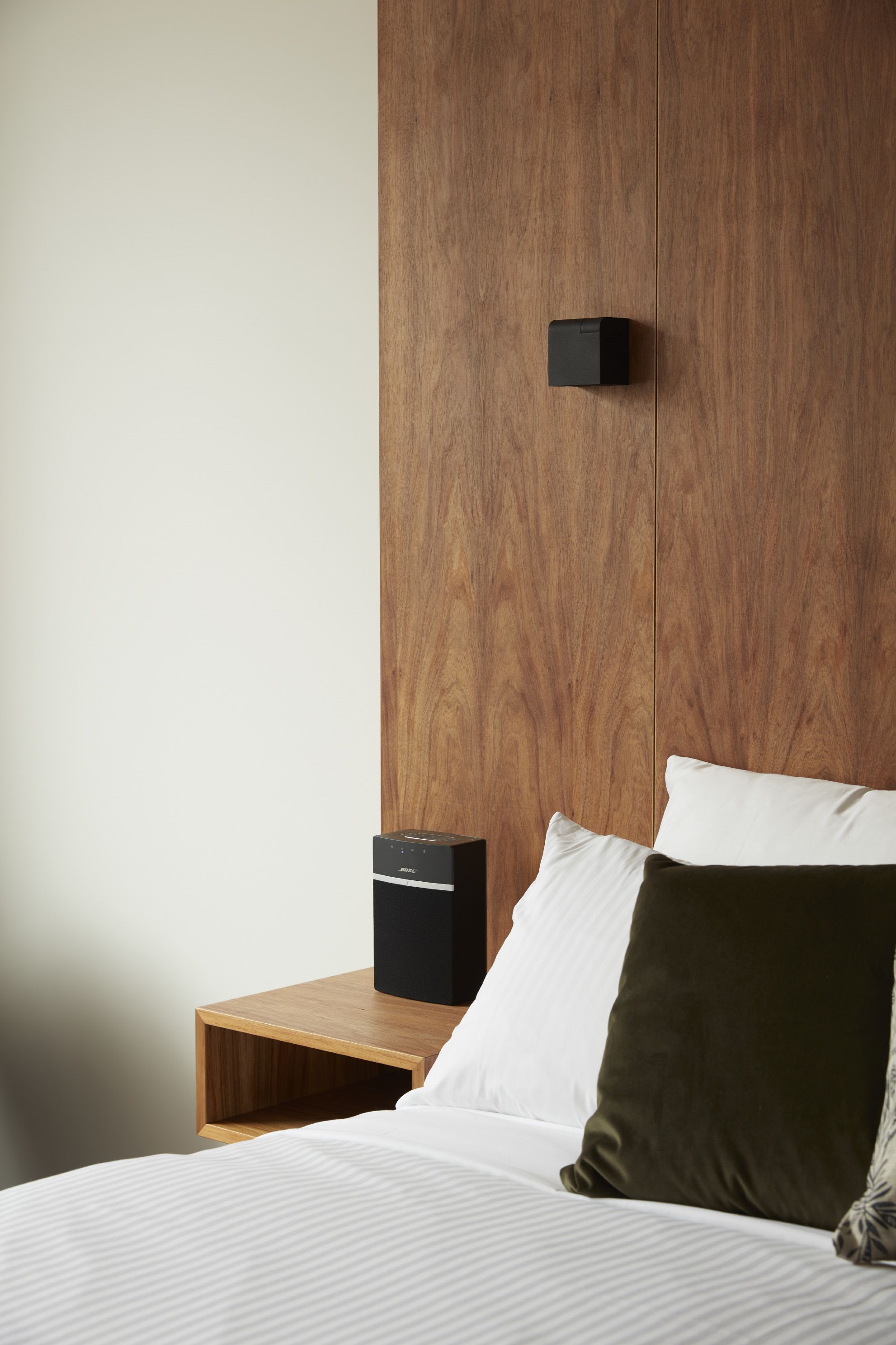 Una cama bien hecha con almohadas blancas y verdes, una mesita de noche de madera y un altavoz negro sobre la mesita.