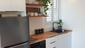 A modern kitchen with white cabinets, a black refrigerator, wooden countertops, a built-in oven, and a window with blinds. - Saltwater Apartments (Lake Cathie)