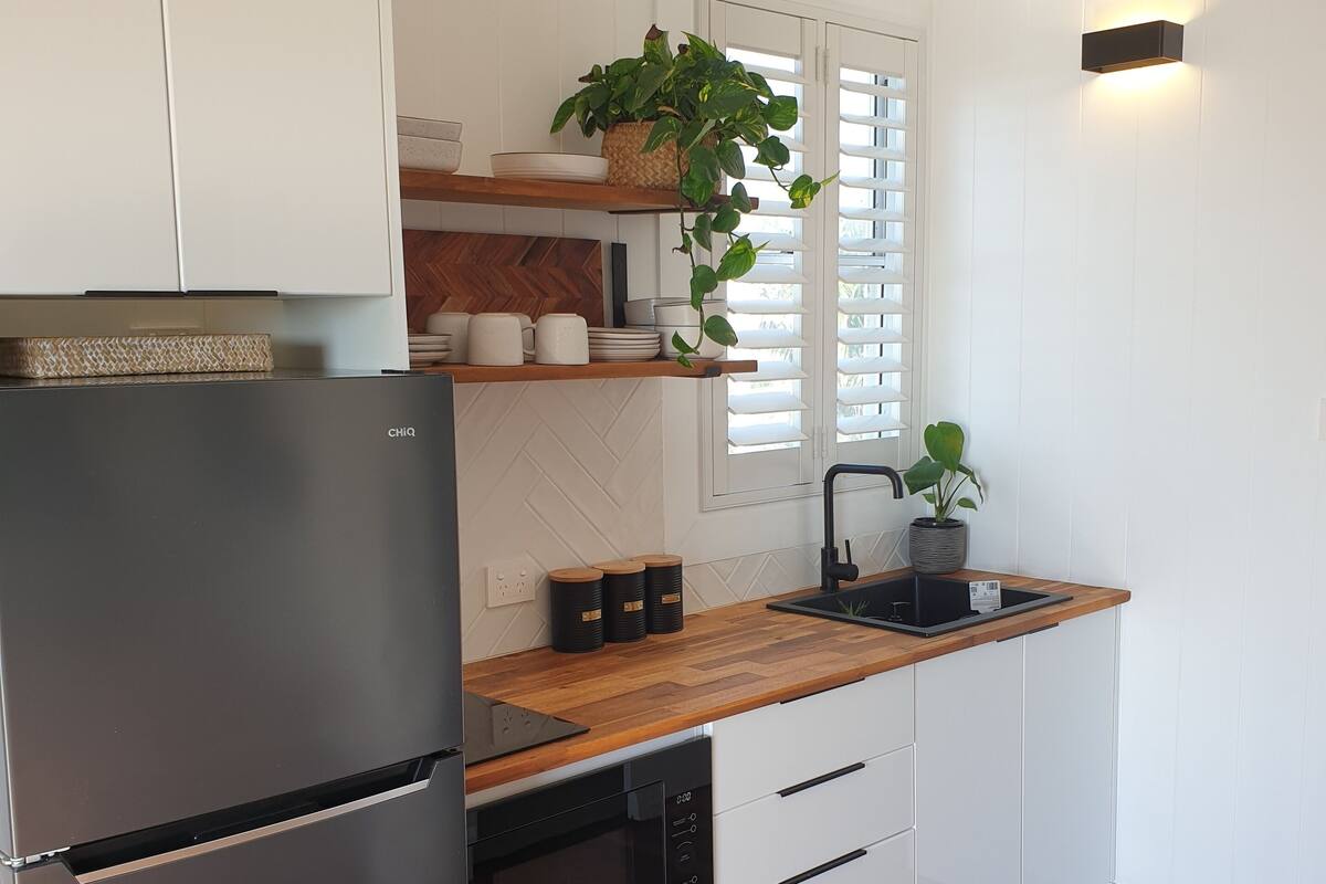 A modern kitchen with white cabinets, a black refrigerator, wooden countertops, a built-in oven, and a window with blinds.