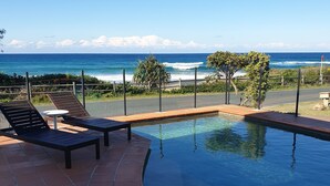 A swimming pool with a curved edge, surrounded by a tiled patio. There are two lounge chairs and a small table. The pool overlooks a beach with the ocean in the background. - Saltwater Apartments (Lake Cathie)