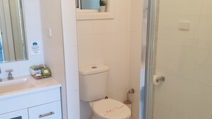 A bathroom with a toilet, a sink with a cabinet, a small window with a plant, and a glass shower enclosure. - Saltwater Apartments (Lake Cathie)