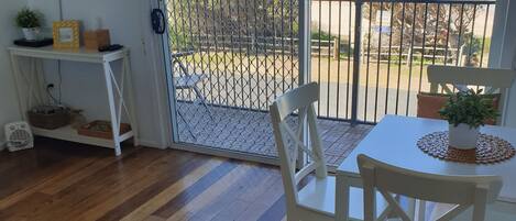 A room with a wooden floor, a white table with two chairs, a TV on the wall, and a view of the ocean through a gate.