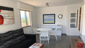 Una sala moderna con un sofá negro, una mesa de comedor blanca con sillas, una mesa de centro con una revista, un cuadro en la pared y vistas al exterior a través de una ventana.