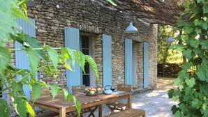 A rustic stone building with a wooden table and benches, a basket of fruit, and a teapot.