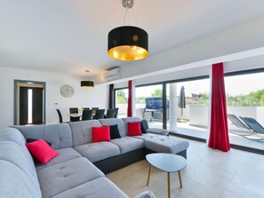 A modern living room with a grey sectional sofa, red cushions, a small round table, and a large window with red curtains. - Vacation home East by Interhome (Vodice/Tribunj)