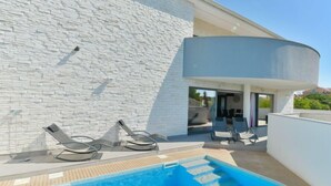 A modern outdoor swimming pool with a stone wall backdrop, lounge chairs, and a curved architectural feature. - Vacation home East by Interhome (Vodice/Tribunj)