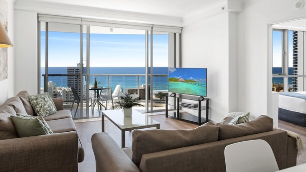 A modern living room with a large TV, comfortable seating, a balcony with a view of the sea, and a dining area. - Chevron Renaissance Ocean View (Surfers Paradise)