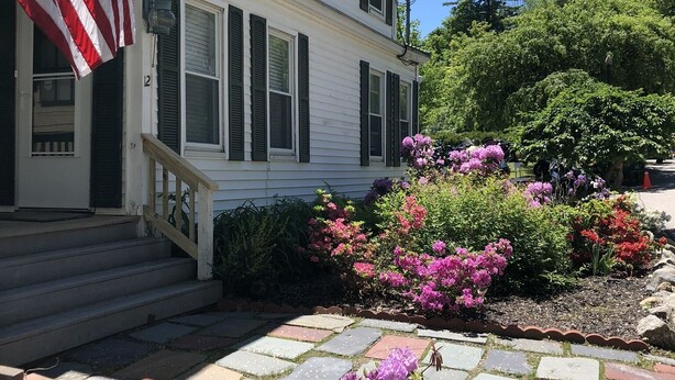 A two-story house with a front porch, a flag, and a garden with flowers. - Only 2 Minutes Walk to Boardwalk, Beach, Eats & Fun! Large Well-Appointed Home. (Weirs Beach, Laconia)