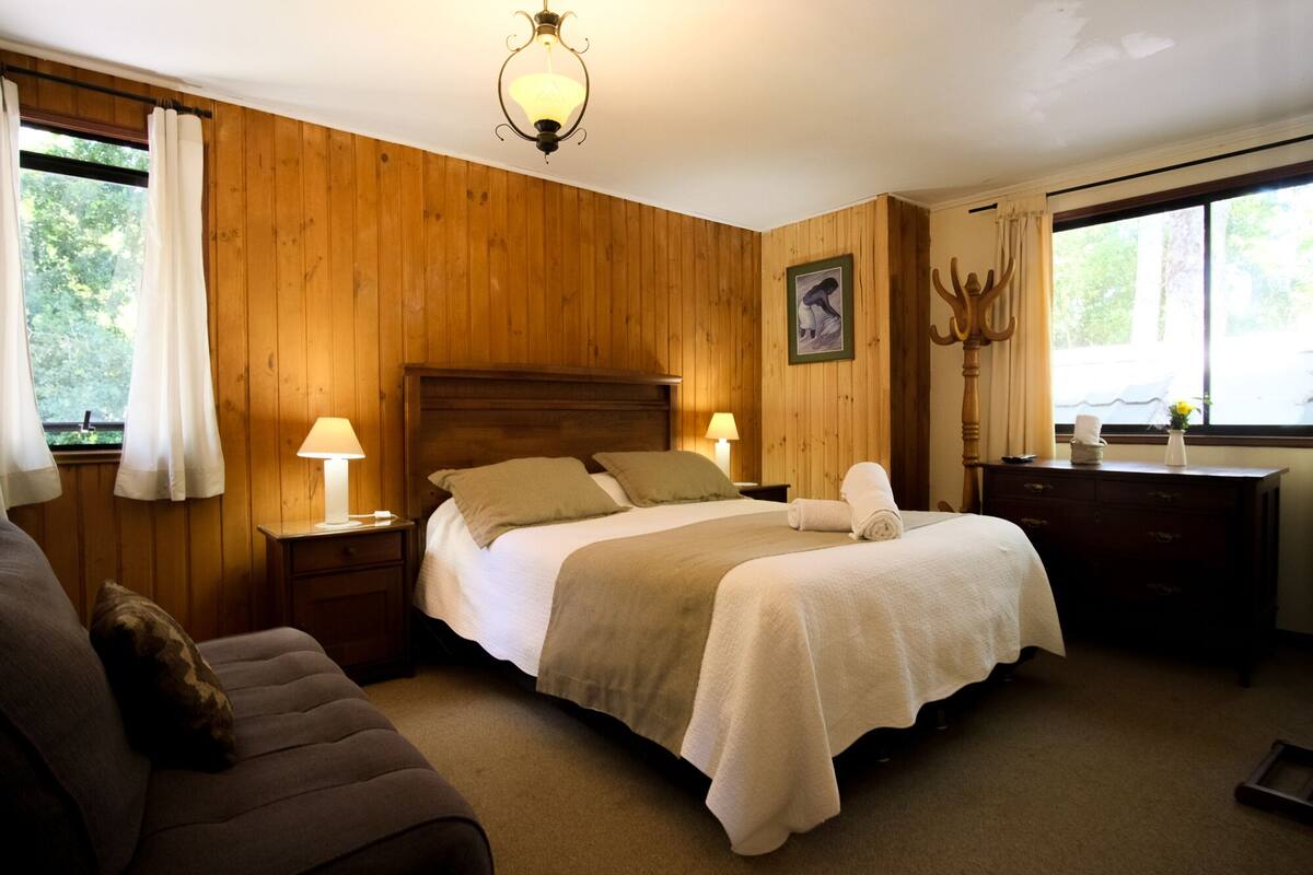 Una habitación acogedora con paredes de madera, una cama con ropa blanca, dos lámparas de noche, un cabecero de madera, un escritorio con una silla y una ventana con cortinas.
