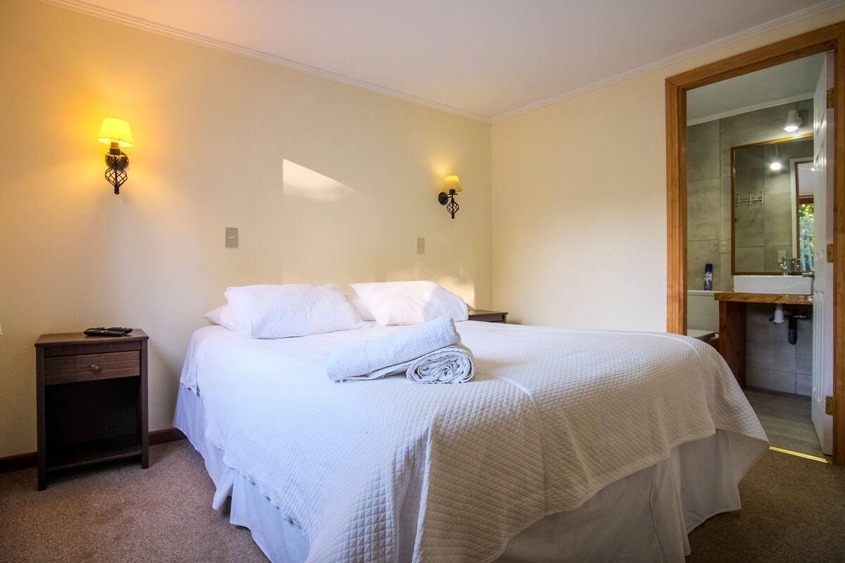 Una cama bien hecha con sábanas blancas, dos mesitas de noche con lámparas, y un espejo grande en el baño.