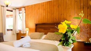 Habitación de hotel con cabecera de madera, cama con ropa blanca, mesita de noche con un jarrón blanco de flores amarillas y una lámpara en la mesita.