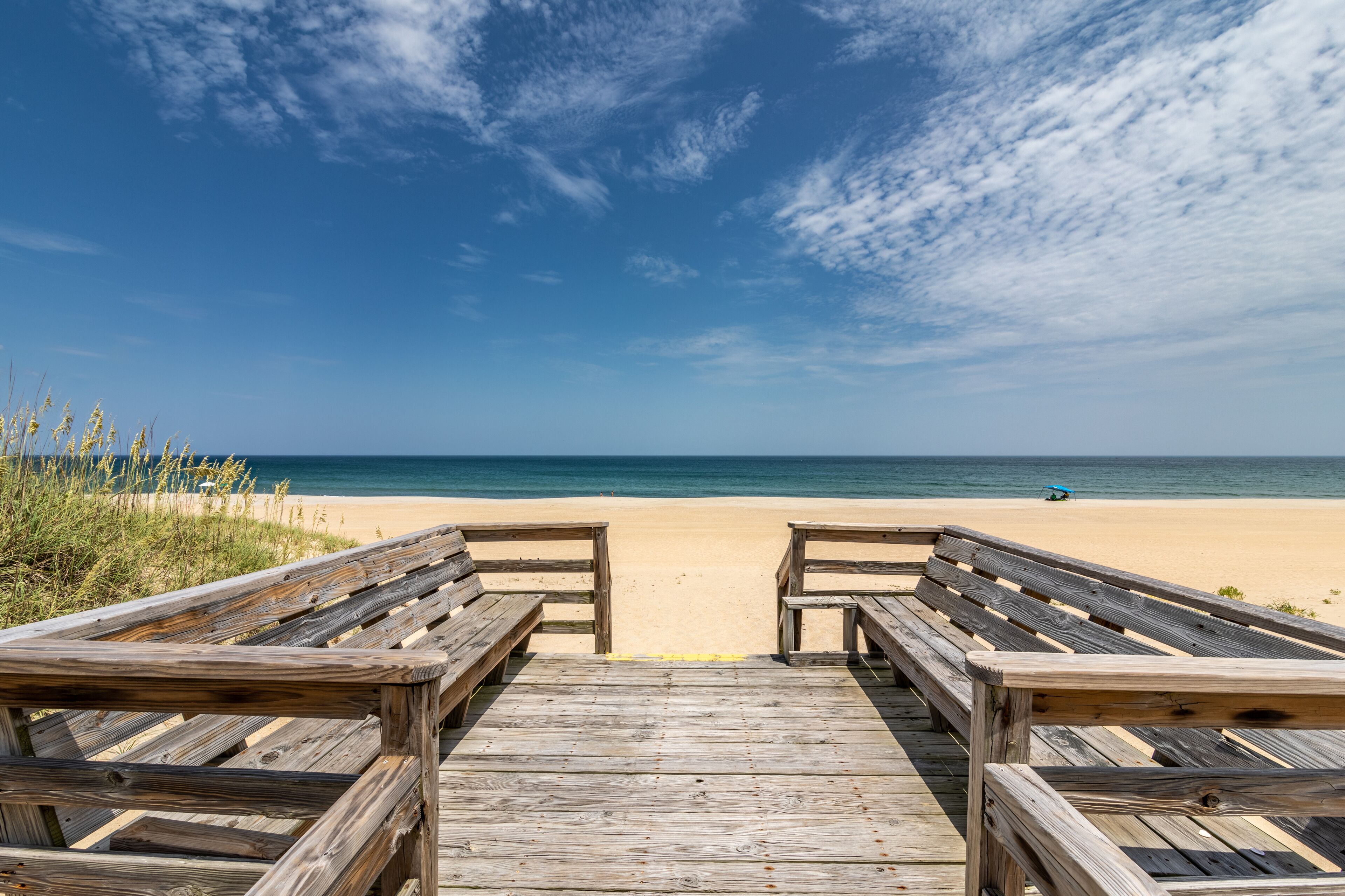 Ein hölzerner Steg führt zu einem Sandstrand, der von einem klaren, blauen Himmel überragt wird, und entfernt ist eine Person mit einem blauen Regenschirm zu sehen.