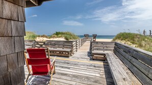 Een houten terras met banken en rode stoelen, dat uitkomt op een strand met duinen en de zee.