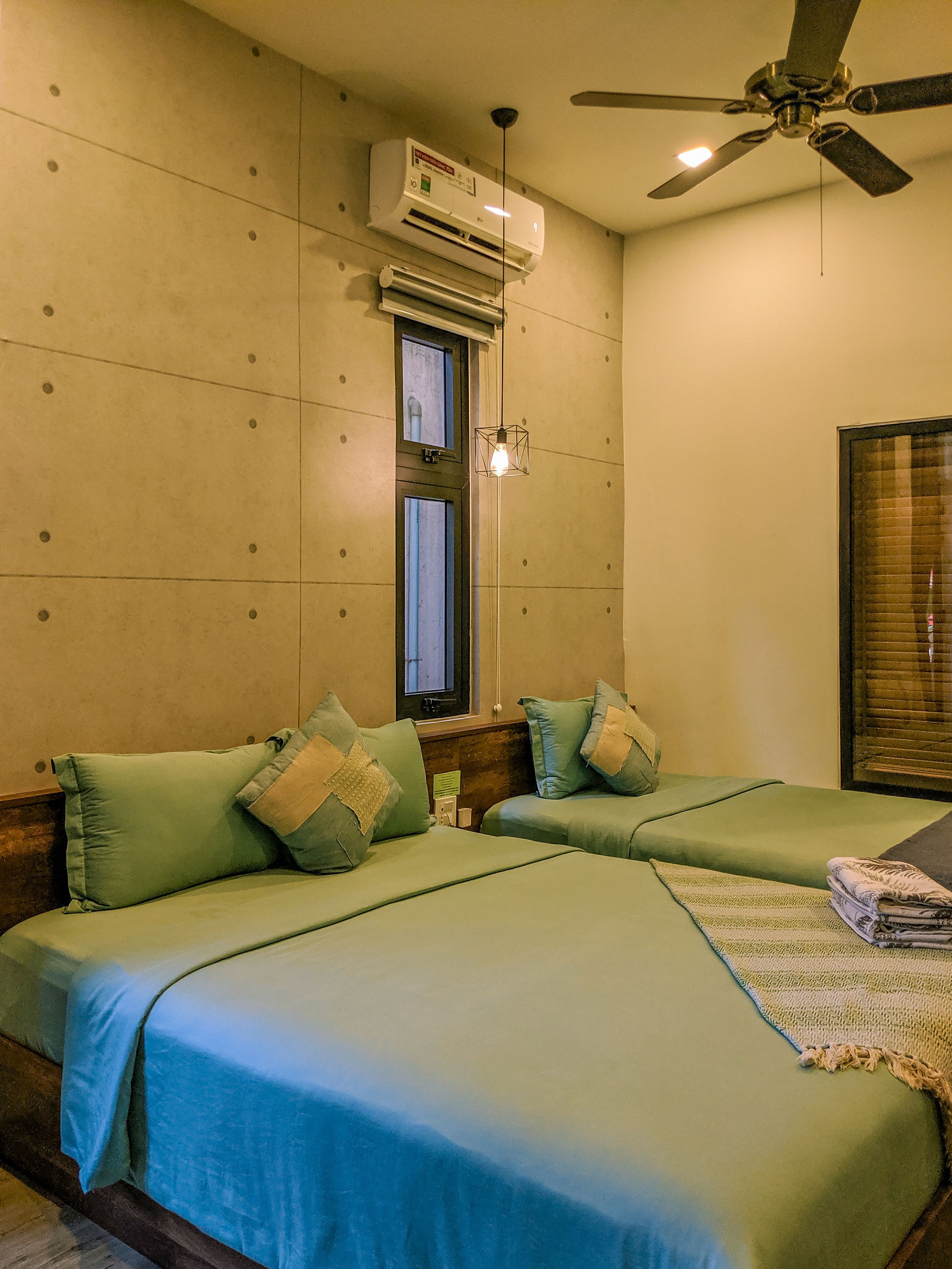 Habitación de hotel con dos camas, unidad de aire acondicionado, ventilador de techo y ventana con persianas.