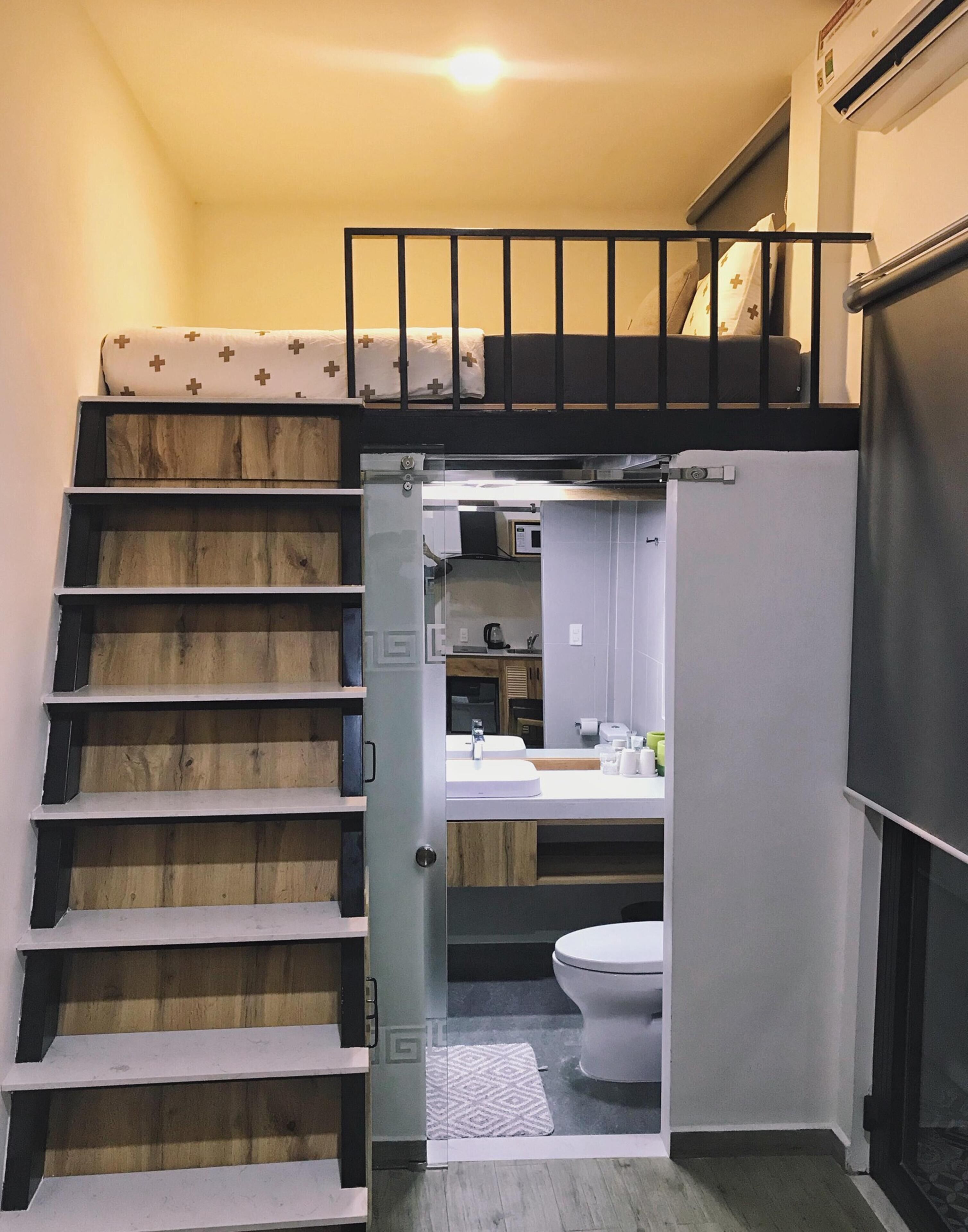 A compact, modern apartment with a small kitchenette, a toilet, and a staircase leading to a loft bed area.
