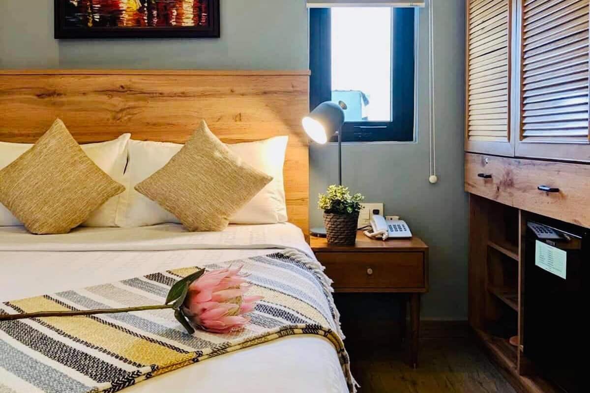 Un dormitorio con cabecera de madera, cama con ropa blanca, mesita de noche con lámpara, ventana y armario de madera.